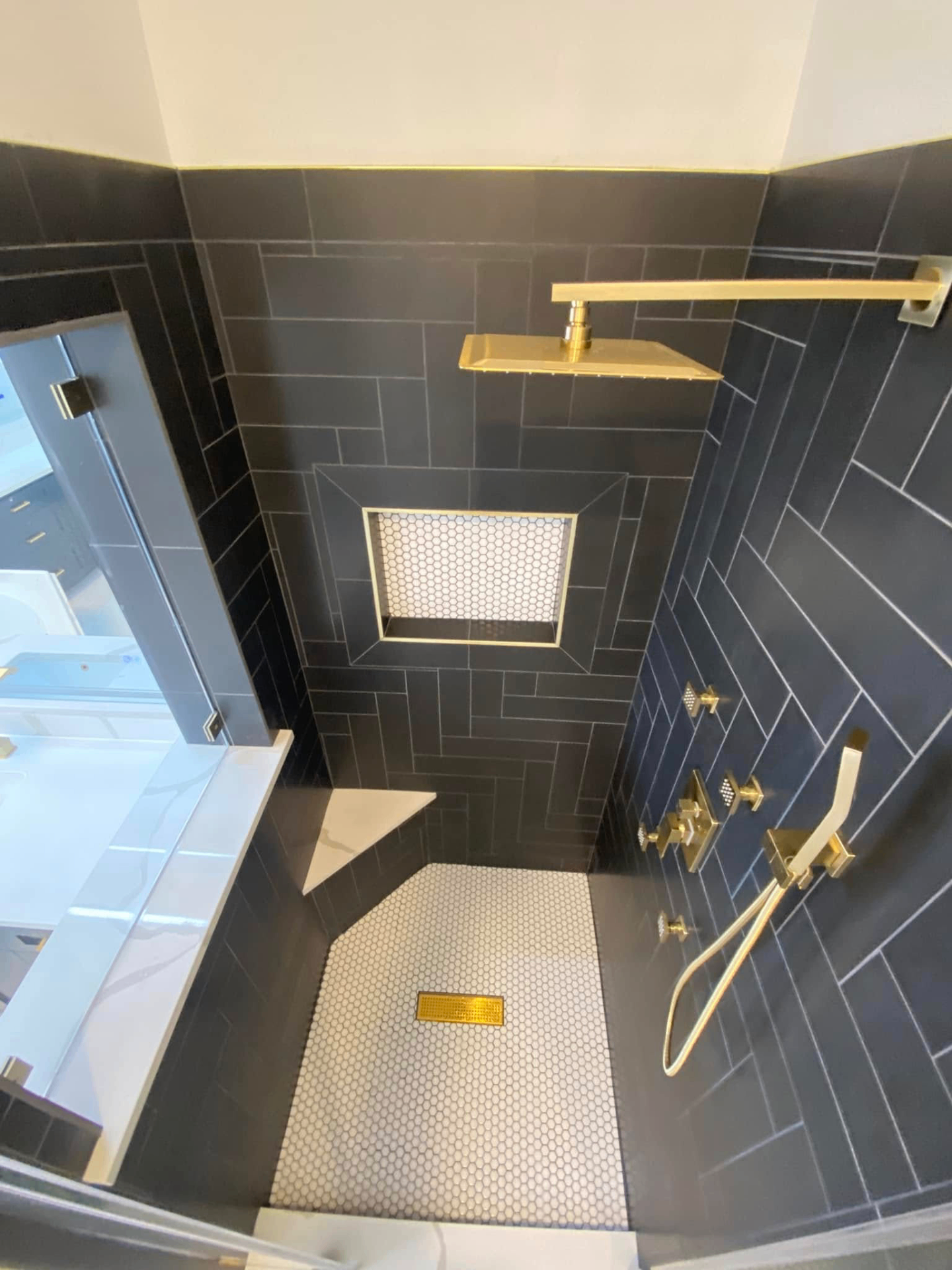 Dark tile walk-in shower with gold fixtures Dark tile walk-in shower with gold rainfall head, body sprays, and hexagon mosaic floor in Richmond TX